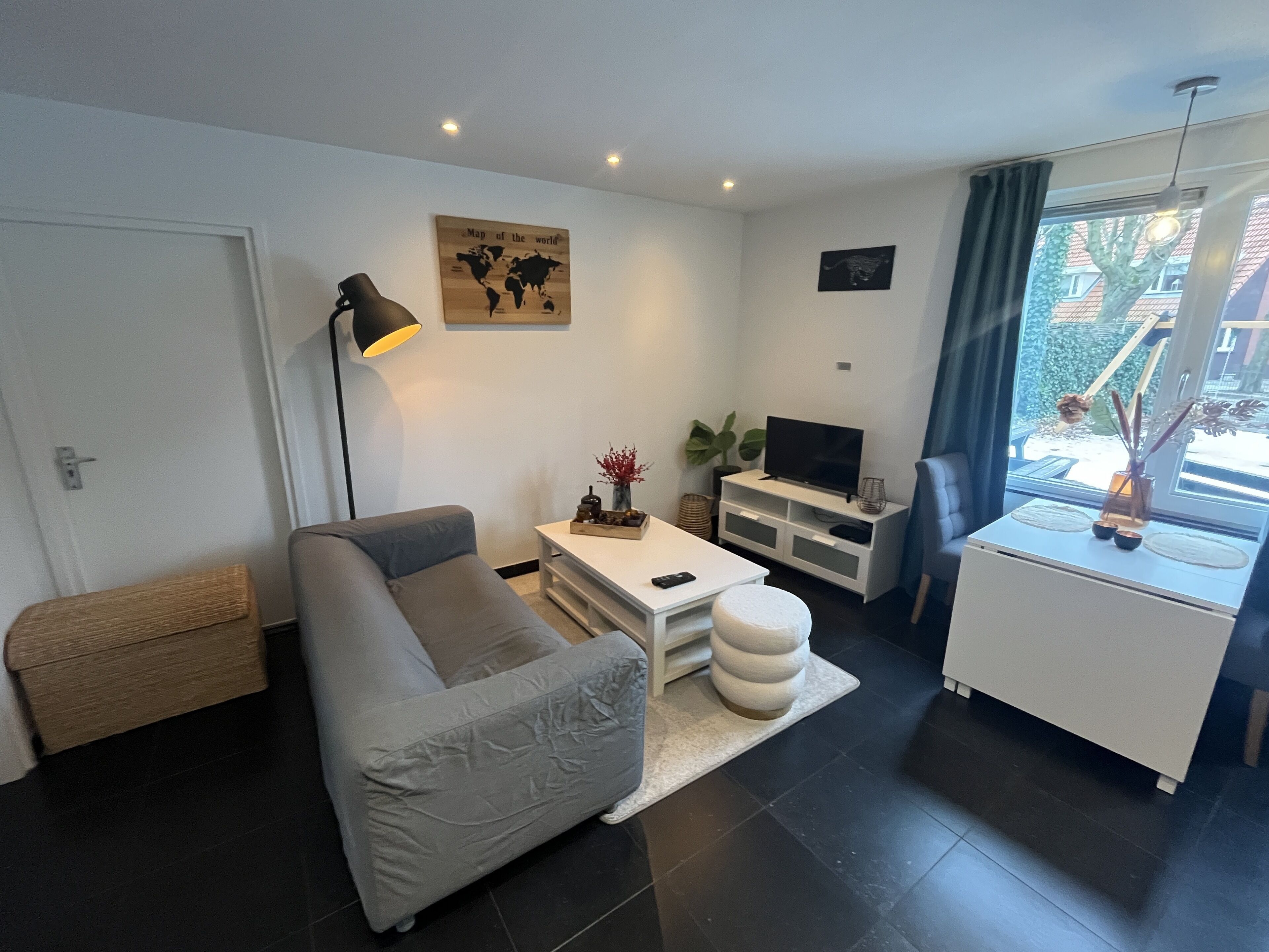 A modern living room with a grey sofa, a coffee table, a TV stand, and a window with a view of a building and greenery outside.