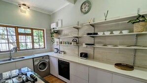 A modern kitchen with a gas stove, washing machine, dishwasher, and shelves with dishes and kitchenware. - Beachside 2BD Home in the Heart of Sea Point! (Cape Town)