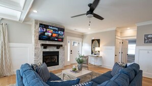 A living room with a stone fireplace, a flat-screen TV mounted above it, and a ceiling fan. - Modern 4BR Oasis - Main Level Master + 2 King Beds (Forest)