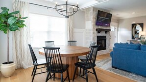 A dining area with a round wooden table and black chairs, a potted plant, and a large window with curtains. - Modern 4BR Oasis - Main Level Master + 2 King Beds (Forest)