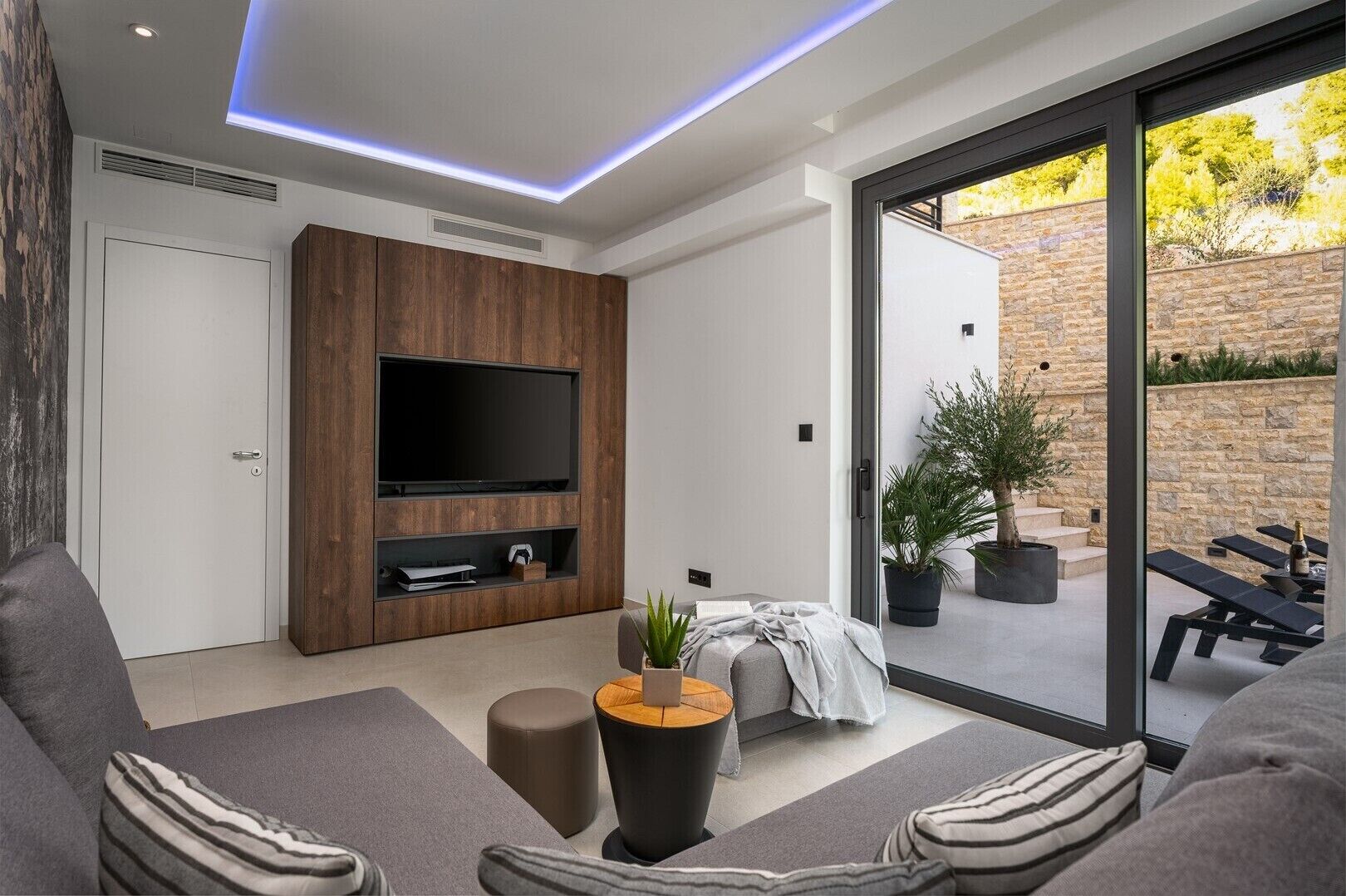Un salon moderne doté d’un téléviseur à écran plat, de canapés gris, d’une table basse et d’une porte-fenêtre coulissante donnant sur un espace extérieur agrémenté de plantes et d’un mur en pierre.