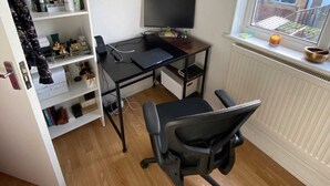 Una habitación pequeña y ordenada con suelo de madera, un escritorio con computadora y silla, un radiador blanco y una ventana con vistas a edificios.