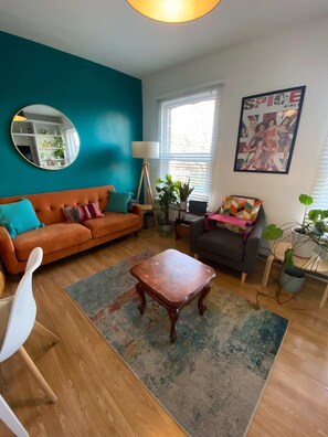 Un salon aux murs de couleur turquoise, avec un canapé marron, une table basse en bois et un fauteuil gris. Un miroir est accroché au mur, ainsi qu’une lampe et une affiche.