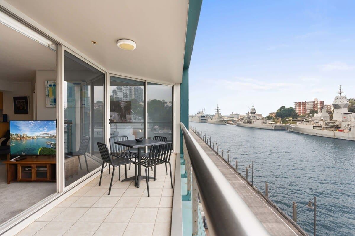 Ein Balkon mit Tisch und Stühlen, mit Blick auf ein Gewässer, im Hintergrund Gebäude und eine Brücke.
