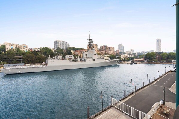 A large naval ship docked at a port with a city skyline in the background. - Apartment in Iconic Fingerwharf (Woolloomooloo)
