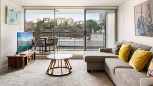A modern living room with a large window, a sofa, a round table, and a painting. - Apartment in Iconic Fingerwharf (Woolloomooloo)