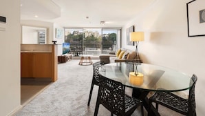 A modern living room with a glass dining table, black chairs, a sofa with cushions, a floor lamp, and a view of buildings through large windows. - Apartment in Iconic Fingerwharf (Woolloomooloo)