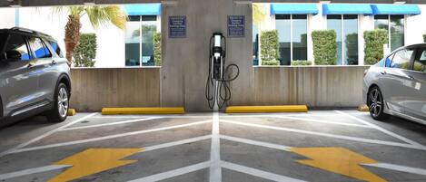 Une borne de recharge pour voitures électriques avec deux voitures garées, un plafond en béton et une vue sur un immeuble doté d’auvents bleus.