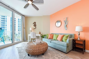 A living room with a large window, a blue sofa, a round wicker coffee table, and a ceiling fan. - Warm Coastal Condo at The Palms with New Floors and Workspace! 🌴 (Destin)