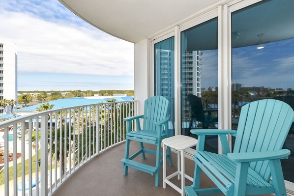 A balcony with two turquoise Adirondack chairs and a small white table, overlooking a pool and a cityscape. - Warm Coastal Condo at The Palms with New Floors and Workspace! 🌴 (Destin)