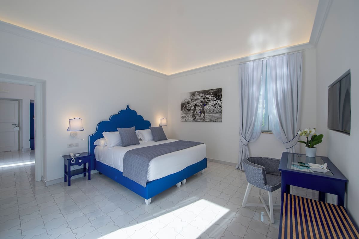 A modern bedroom with a blue bed, a desk with a chair, a small table with a lamp, and a large window with curtains.