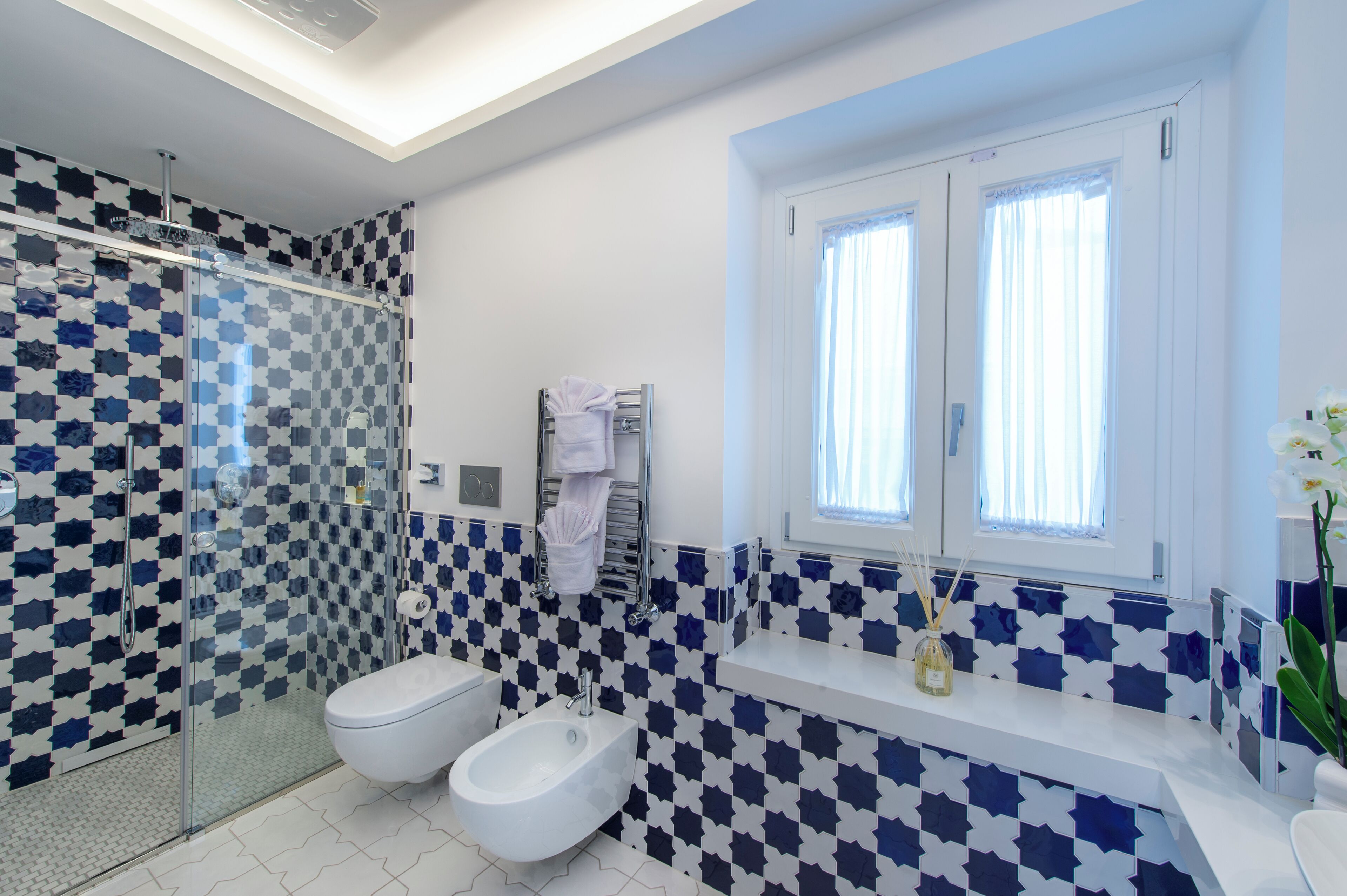 Un bagno moderno con zoccolo a scacchi, cabina doccia in vetro, bidet e finestra con tende.