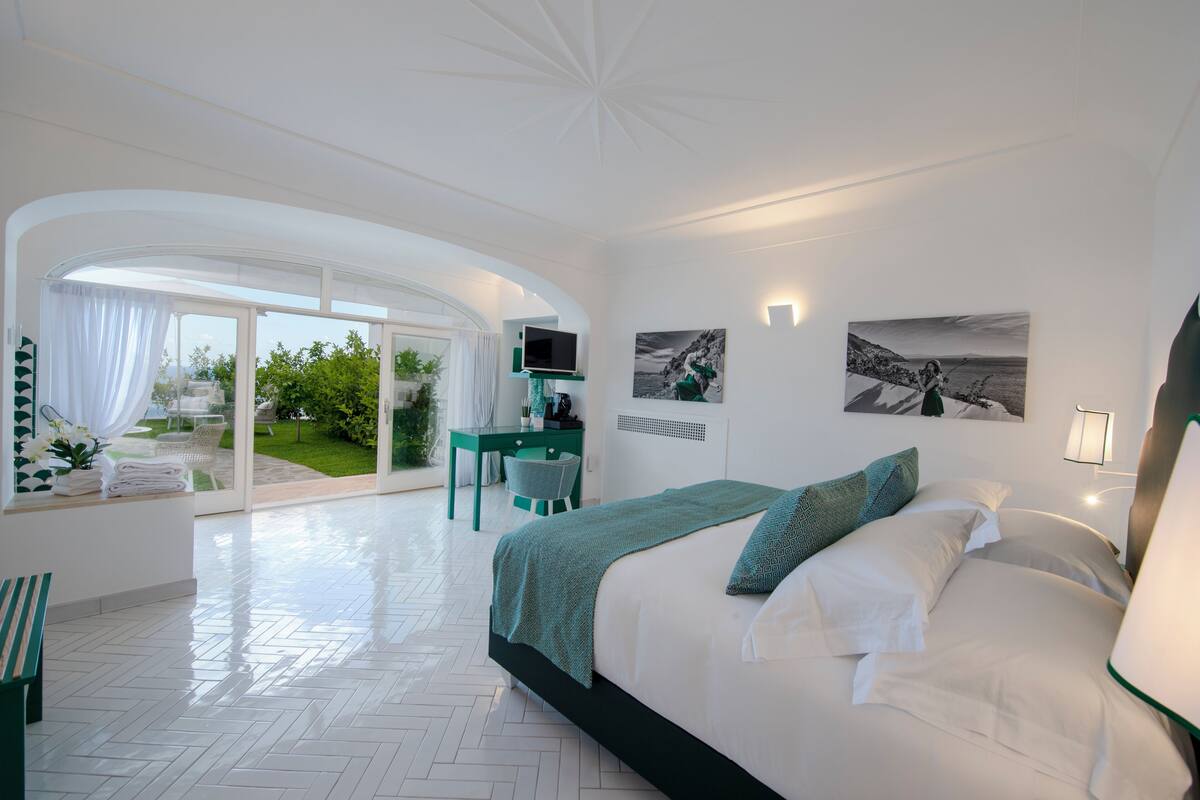 A modern hotel room with a large bed, a desk, and a view of a garden.