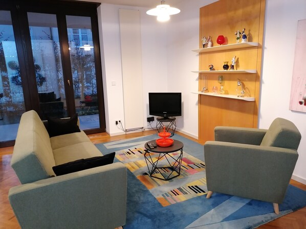 A modern living room with two grey sofas, a flat-screen TV on a stand, a small coffee table with a red bowl, and a colorful rug.