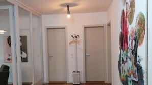 Een hal met houten vloer, witte muren en deuren. Er staat een kapstok met hangers, een ventilatie rooster en een plafondlamp.