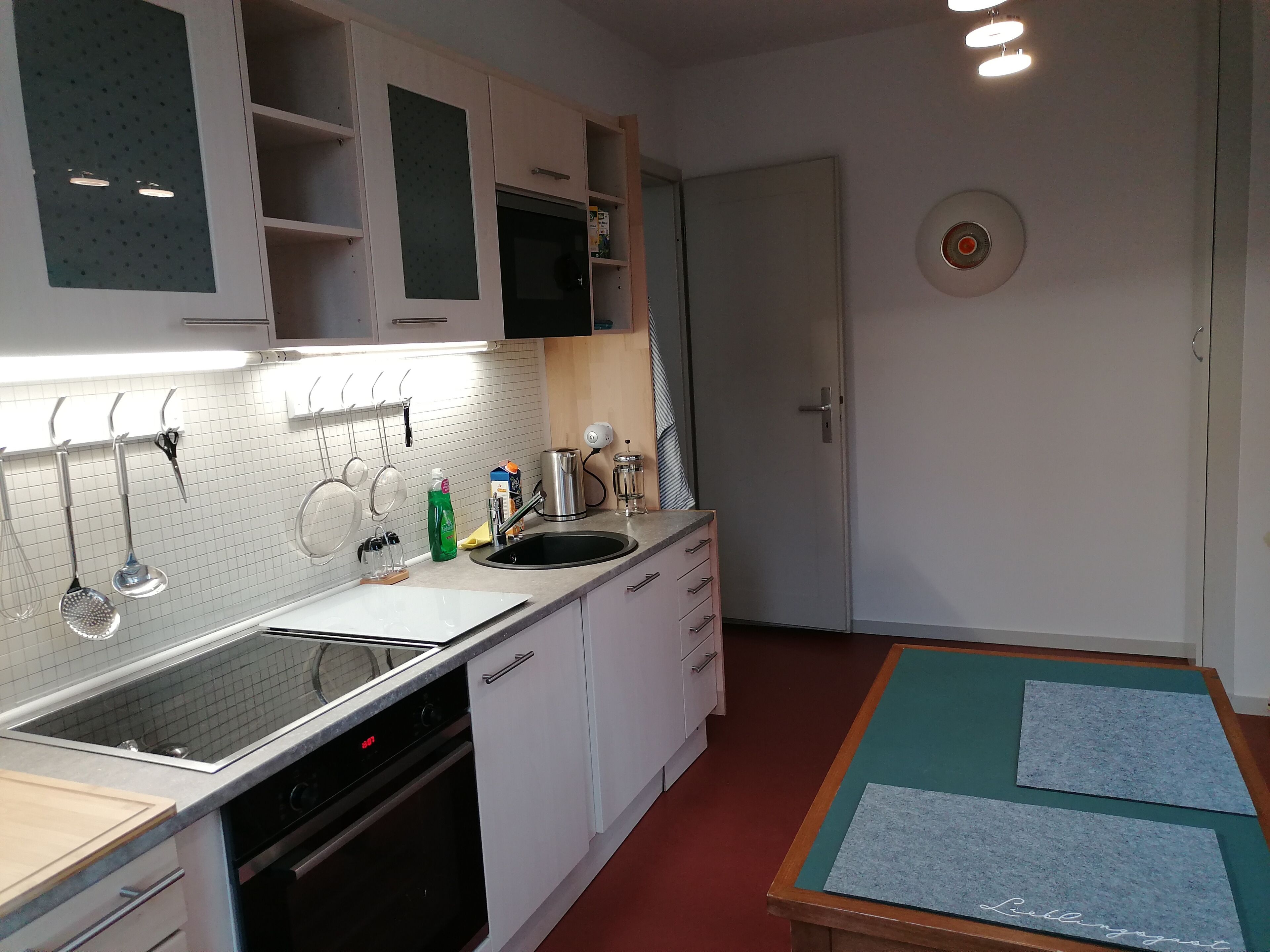 Une cuisine moderne avec des meubles blancs, un évier noir et des appareils en acier inoxydable. On y trouve une table en bois, recouverte d’une nappe verte, et une plaque nominative.