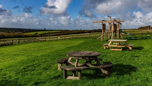 A picnic area with wooden tables and benches, a swing set, and a view of the countryside. - The Linhay, shared hot tub and sauna (Hartland)