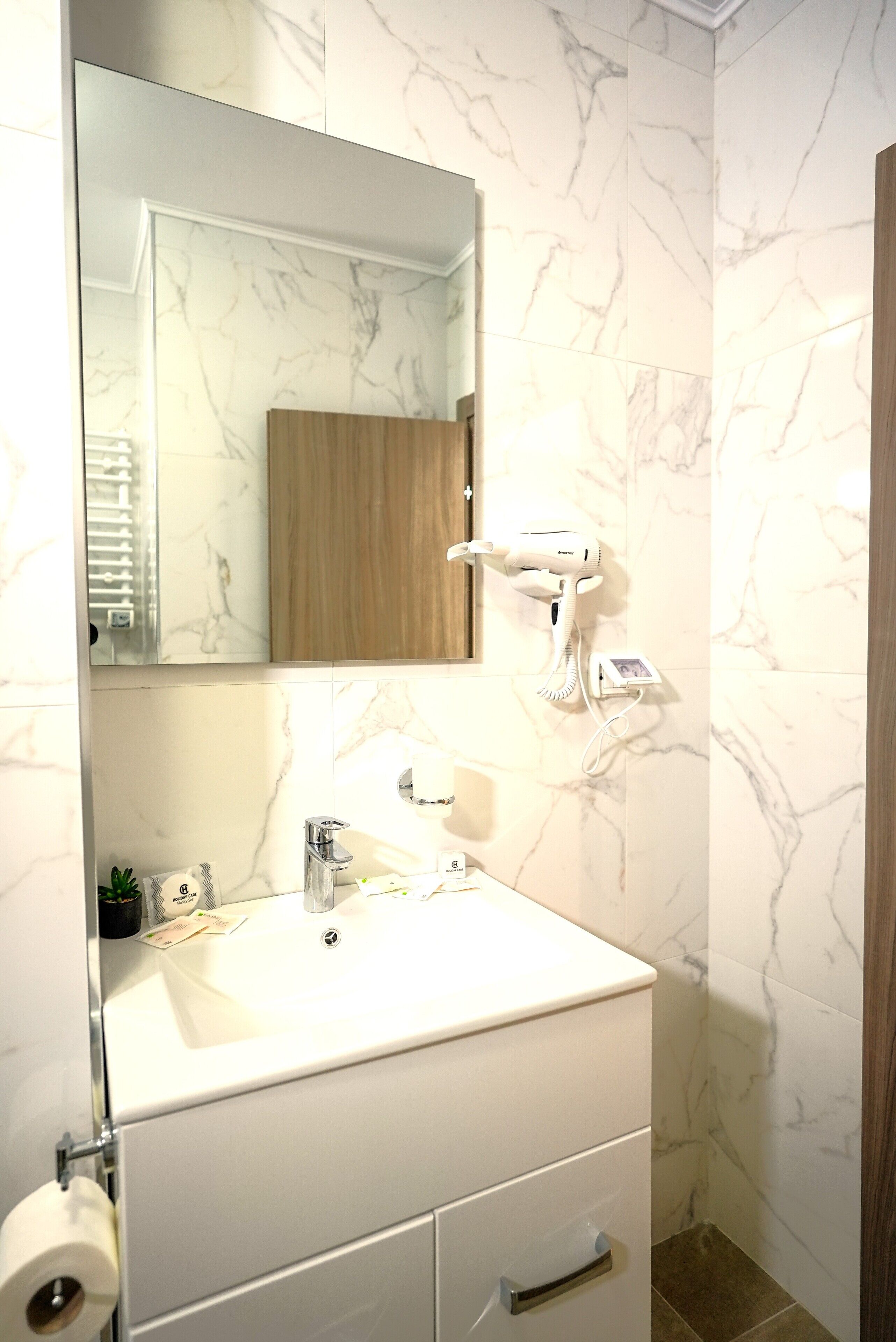 A bathroom with a marble wall, a wooden cabinet, a white sink, and a hairdryer mounted on the wall.