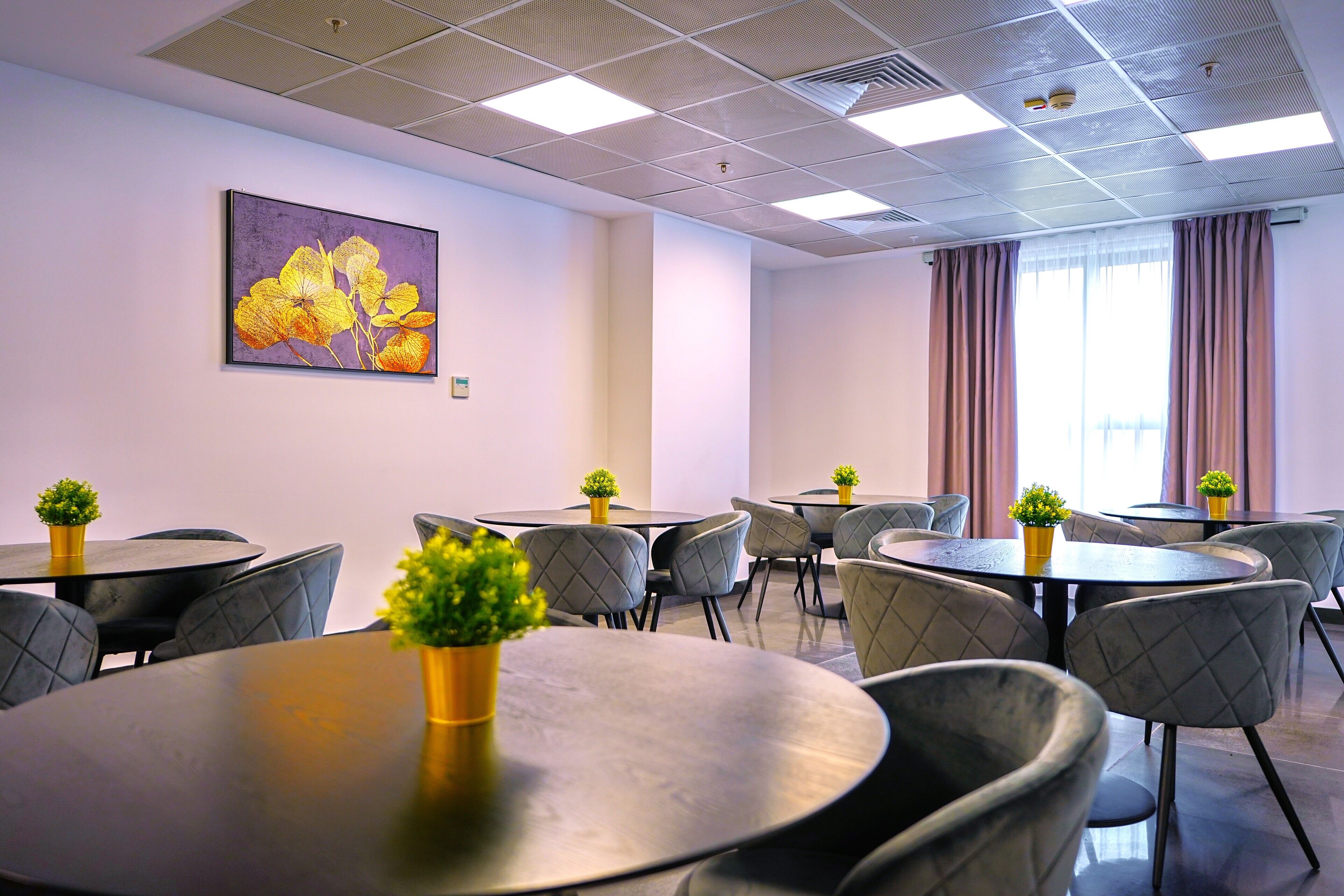 A modern hotel room with round tables and grey upholstered chairs, a painting on the wall, and a large window with curtains.