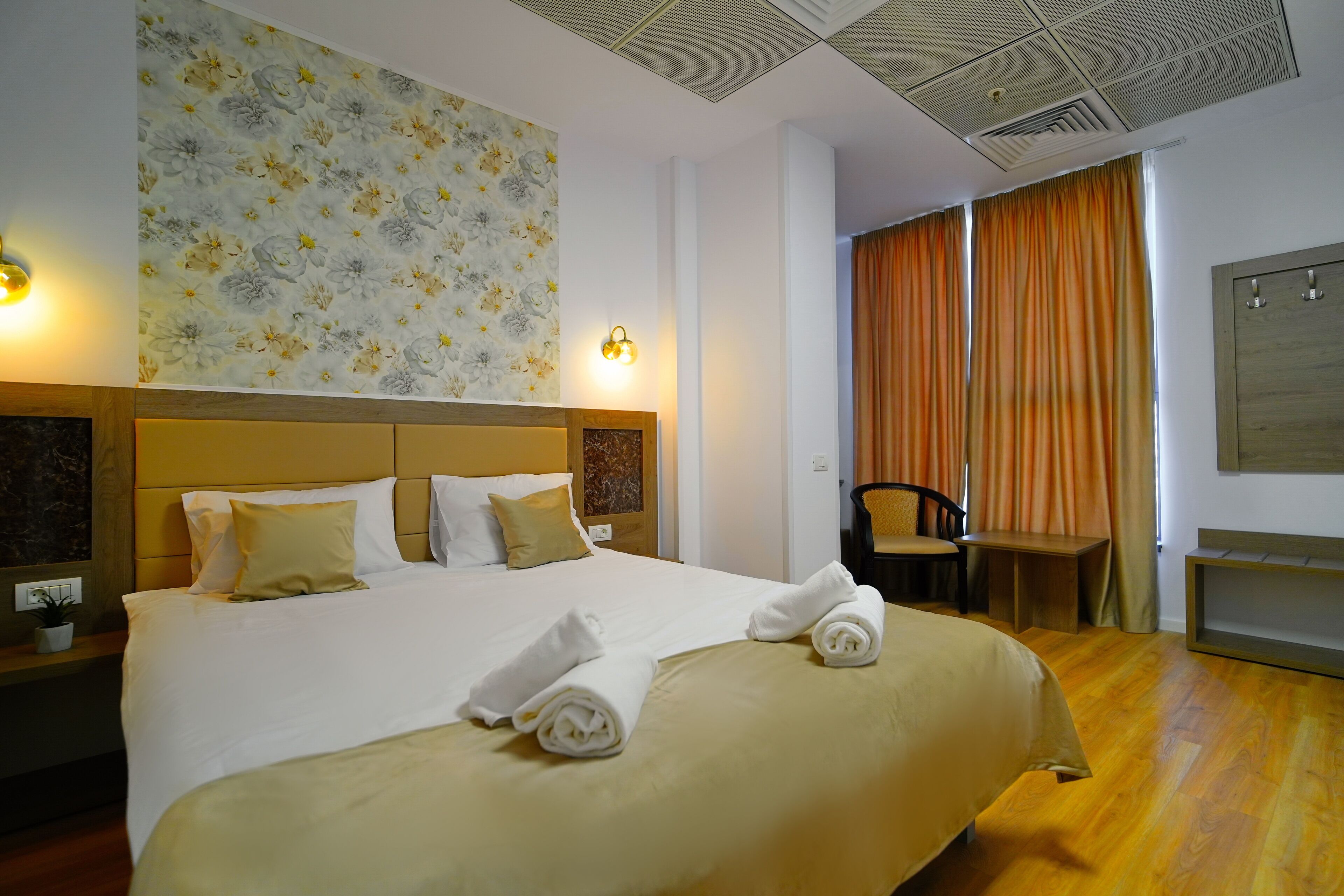 A hotel room with a large bed, a headboard, a wall-mounted light, a desk, a chair, and a window with curtains.
