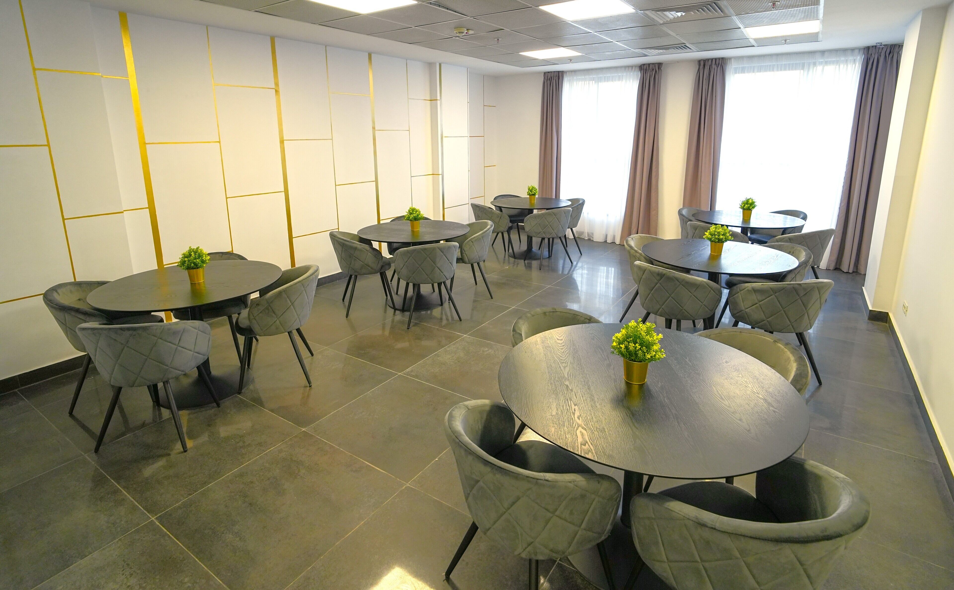 Un salón de conferencias con varias mesas redondas y sillas, con un diseño moderno que incluye paneles de pared geométricos y amplios ventanales con cortinas.