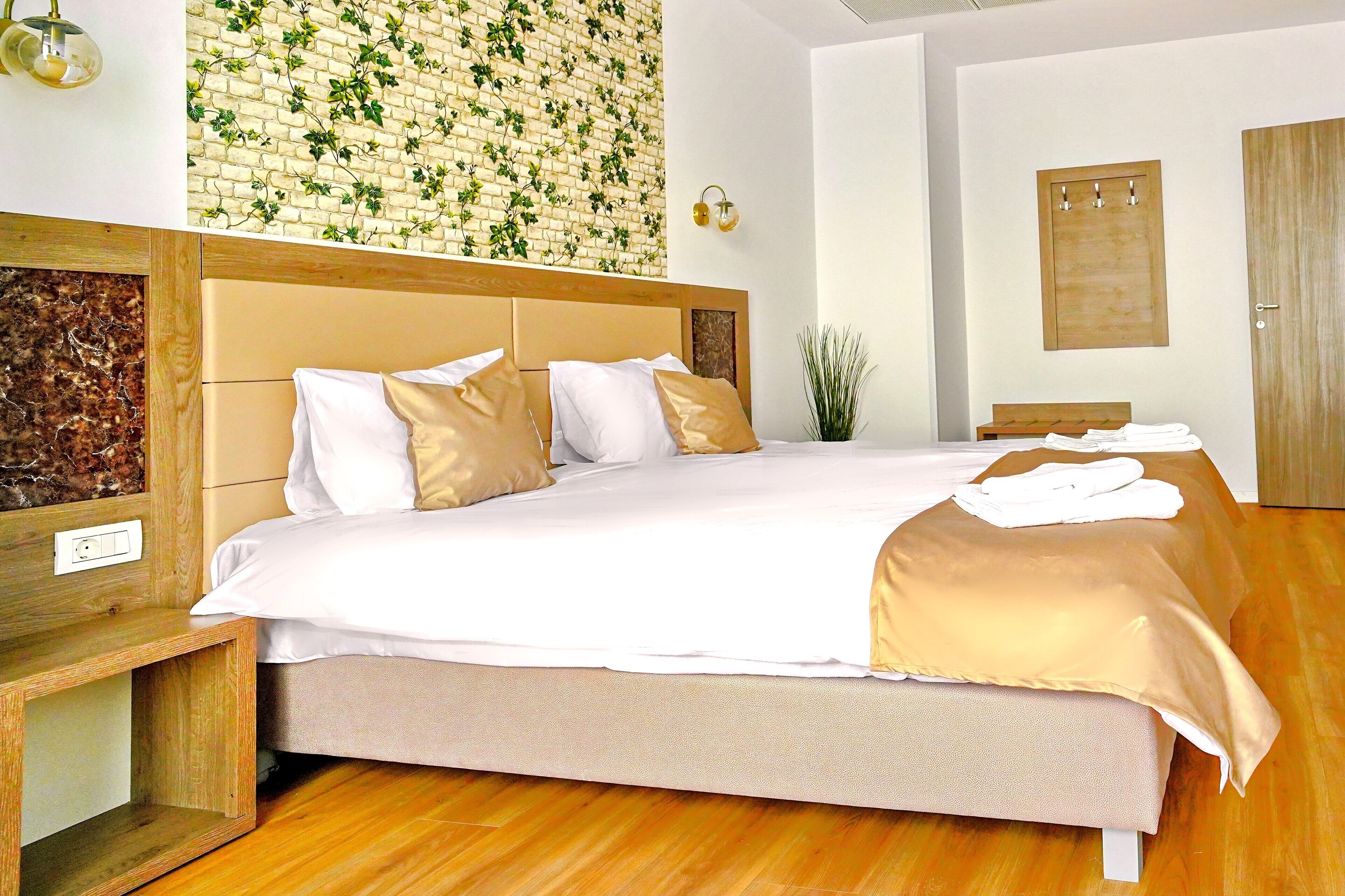 A neatly made bed with white and beige bedding, a wooden headboard, and a wall with a brick pattern and green plants.