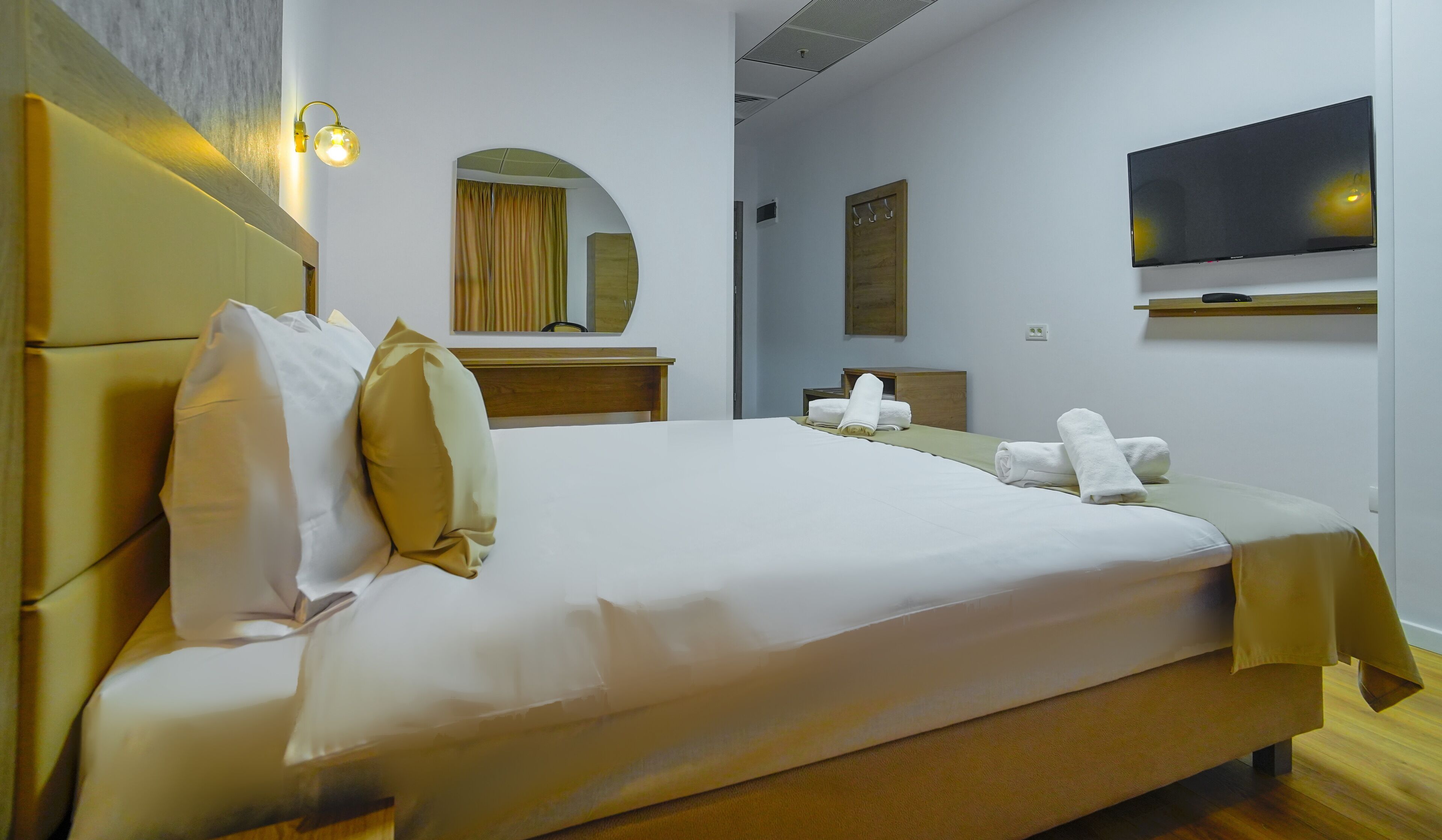 A hotel room with a large bed, a wooden headboard, a flat-screen TV mounted on the wall, and a wooden nightstand with a mirror.