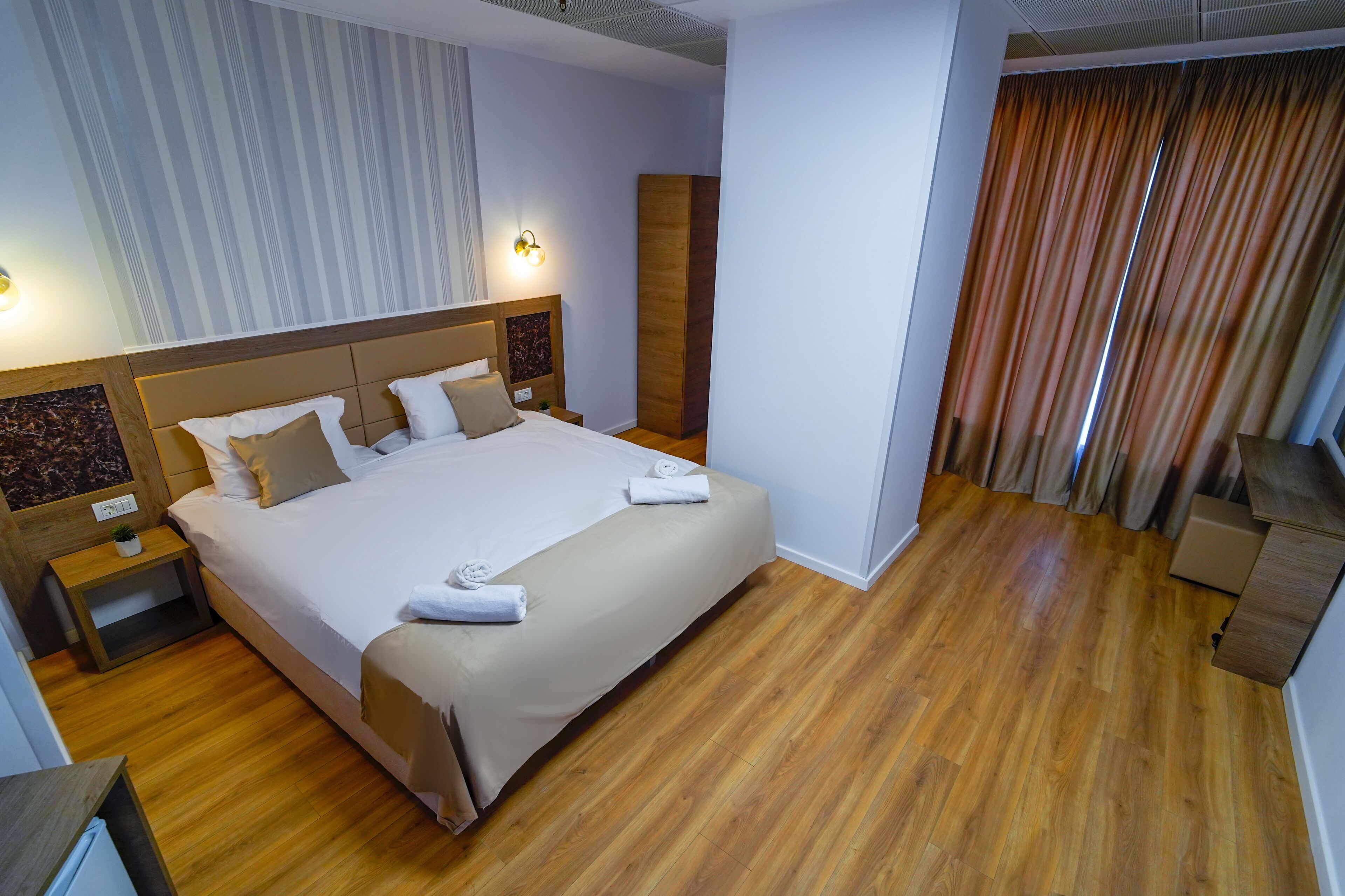 Habitación de hotel con una cama grande, suelo de madera, una mesita de noche, una lámpara de pared y una cortina.