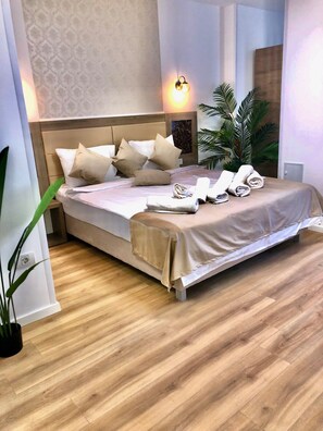 A neatly made bed with beige linens and pillows, a bedside table with a lamp, and a potted plant beside the bed. - Sir Fundeni Hotel (Bucharest)