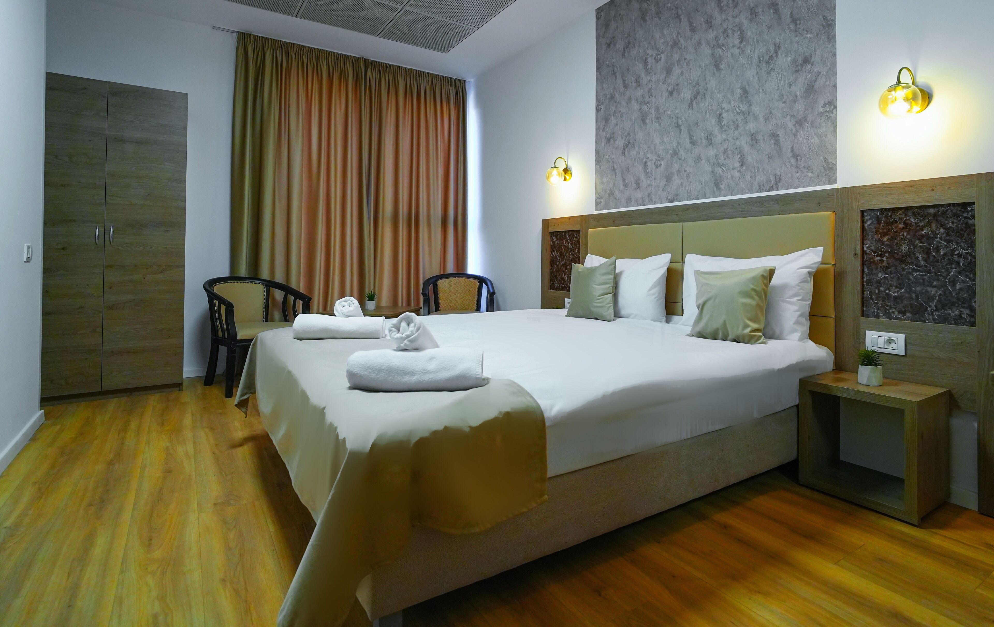 A hotel room with a large bed, two chairs, a nightstand, and a wall-mounted light fixture.