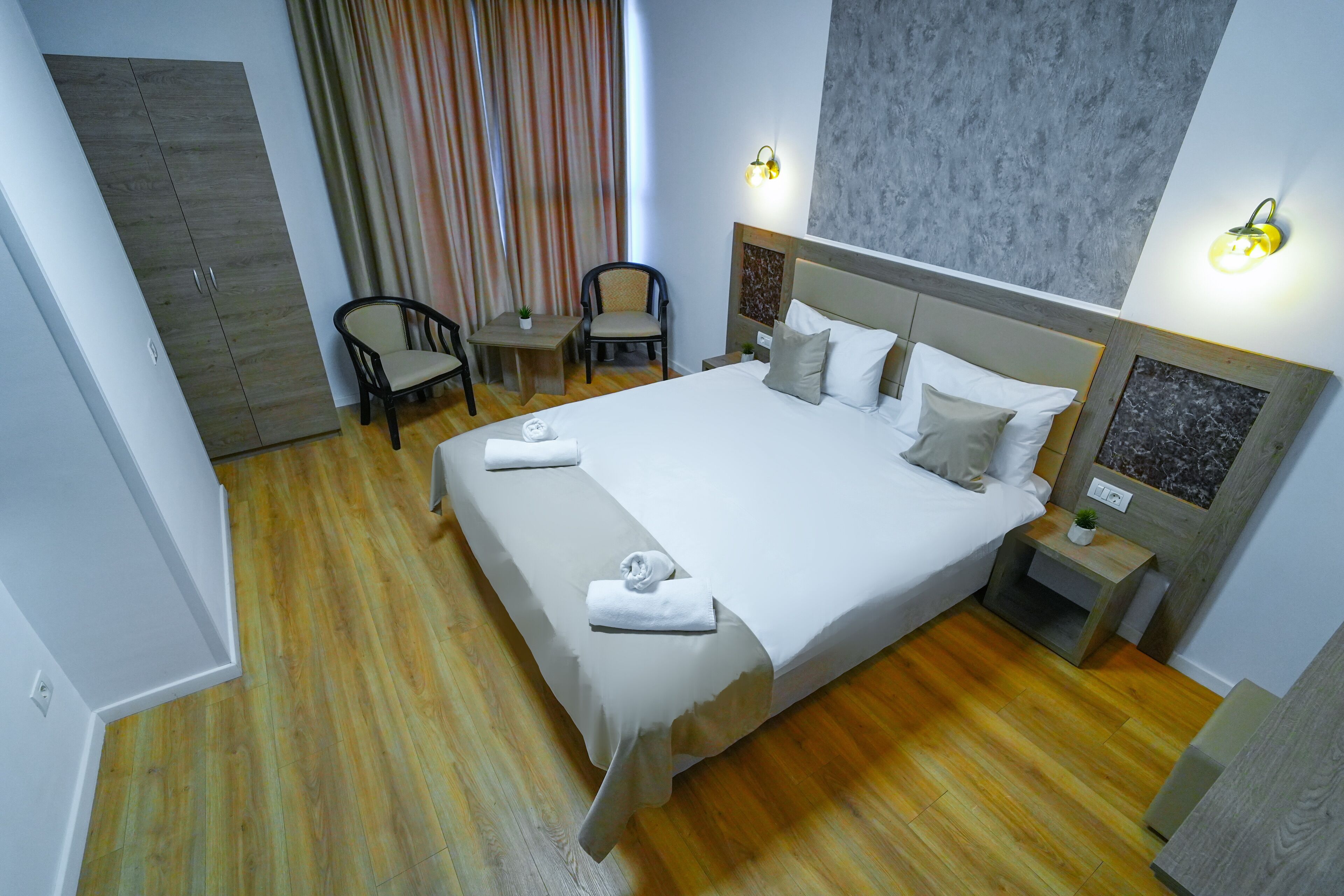 Una habitación de hotel con una cama grande, dos mesitas de noche, una silla, una pequeña mesa y un armario.