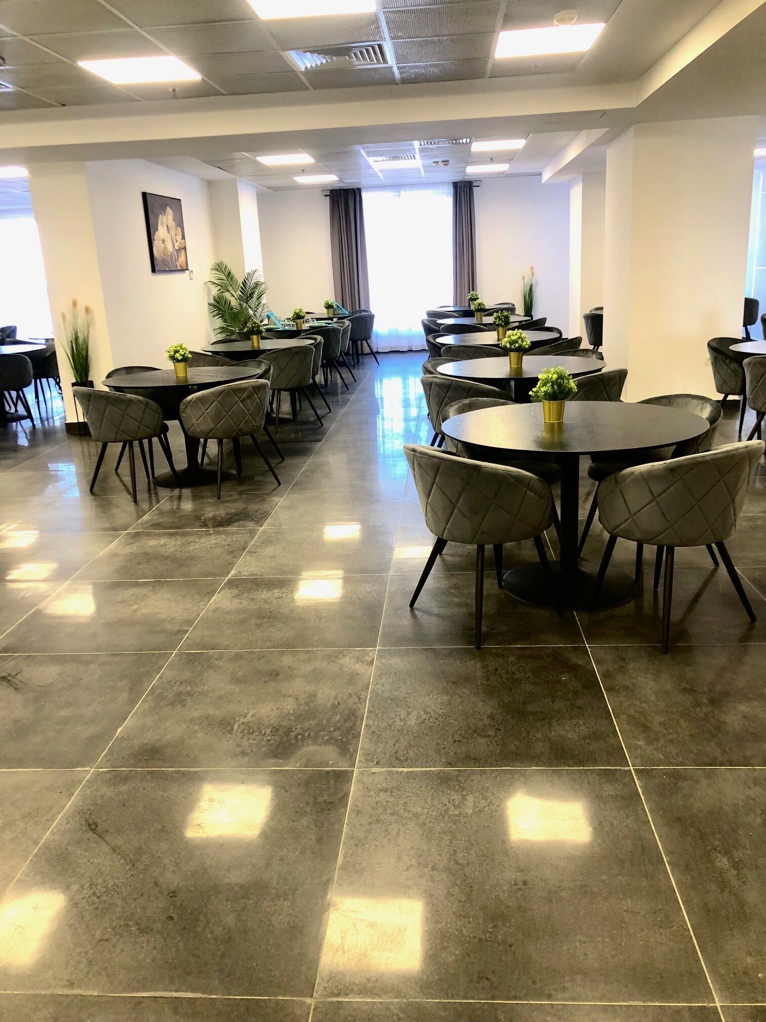 Een ruime vergaderzaal met ronde tafels en stoelen, grote ramen met gordijnen en potplanten.