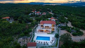 A large house with a swimming pool, surrounded by dense greenery and a scenic sunset view.
