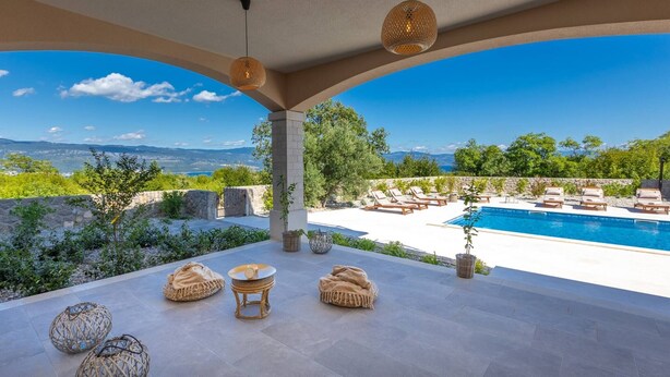 A poolside area with lounge chairs, a table, and a view of the sea and mountains.