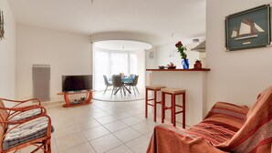 Un salon équipé d’un téléviseur, de meubles en osier, d’un coin repas avec des chaises et une table, ainsi que d’un coin cuisine avec une étagère et un tableau.