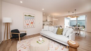 A modern living room with a white sofa, a black armchair, and a round coffee table. There is a painting on the wall, a dining area with a table and chairs, and a kitchen in the background. - Barefoot Beach House McCrae (McCrae)