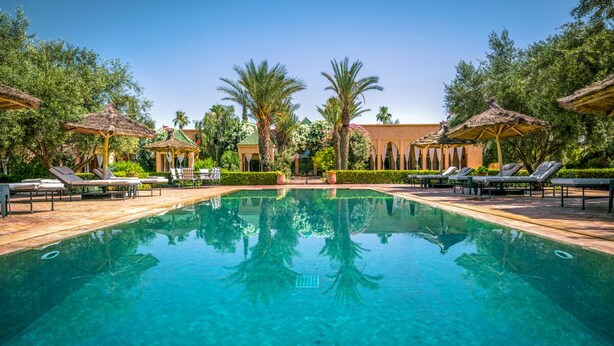 A swimming pool with lounge chairs and umbrellas, surrounded by greenery and a building in the background. - Villa Tounsi - Five Bedroom Villa, Sleeps 10 (Douar Tounsi)
