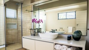 Un baño moderno con ducha envuelta en vidrio, un espejo grande, un lavamanos blanco y un mueble de tocador con toallas.