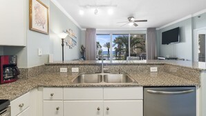 A modern kitchen with granite countertops, a double sink, and a dishwasher. There is a ceiling fan, a flat-screen TV, and a view of palm trees and a pool through large windows.