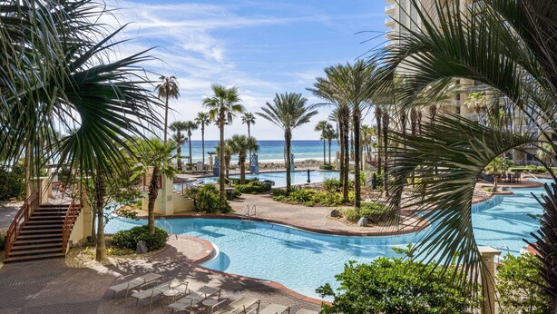 A resort pool area with palm trees, a view of the ocean, and a building in the background.