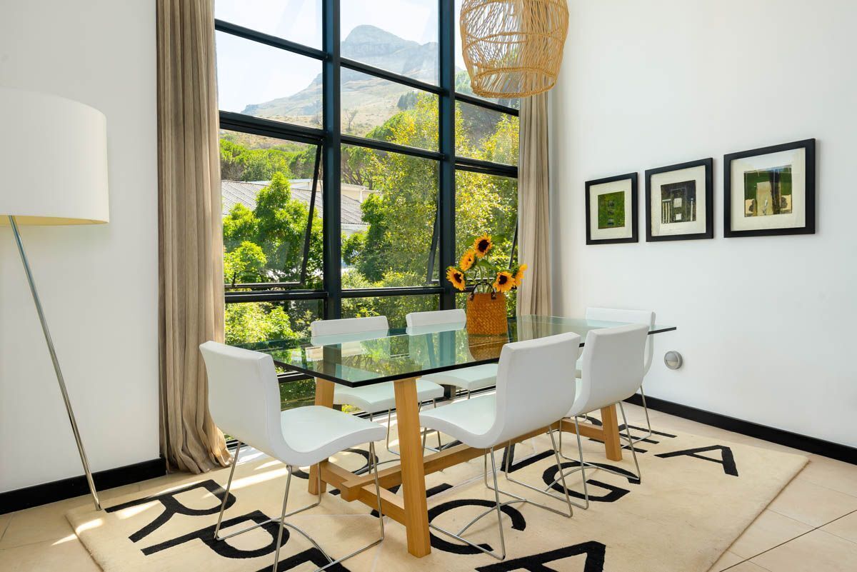 Un comedor moderno con una mesa de vidrio, sillas blancas y vistas a la vegetación a través de amplios ventanales.