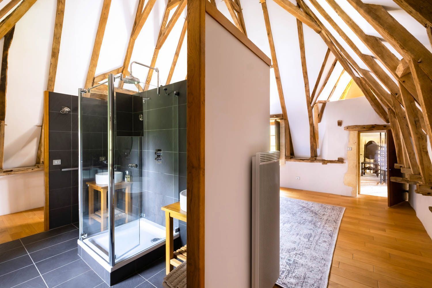 A modern bathroom with a glass shower enclosure, dark tiles, and wooden beams in the ceiling.