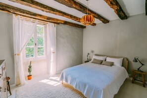 Un dormitorio con cama, mesita de noche, lámpara, ventana con cortinas y una planta de cactus.