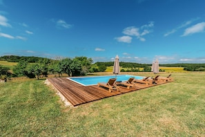 A rectangular swimming pool with a wooden deck, surrounded by lounge chairs and umbrellas, set against a rural landscape.
