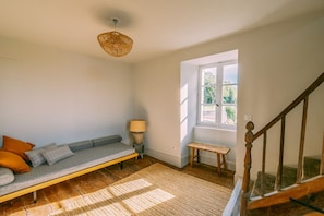 Una habitación con piso de madera, un sofá gris con cojines, una lámpara de techo tejida, una mesita con lámpara y una ventana con vistas a follaje.