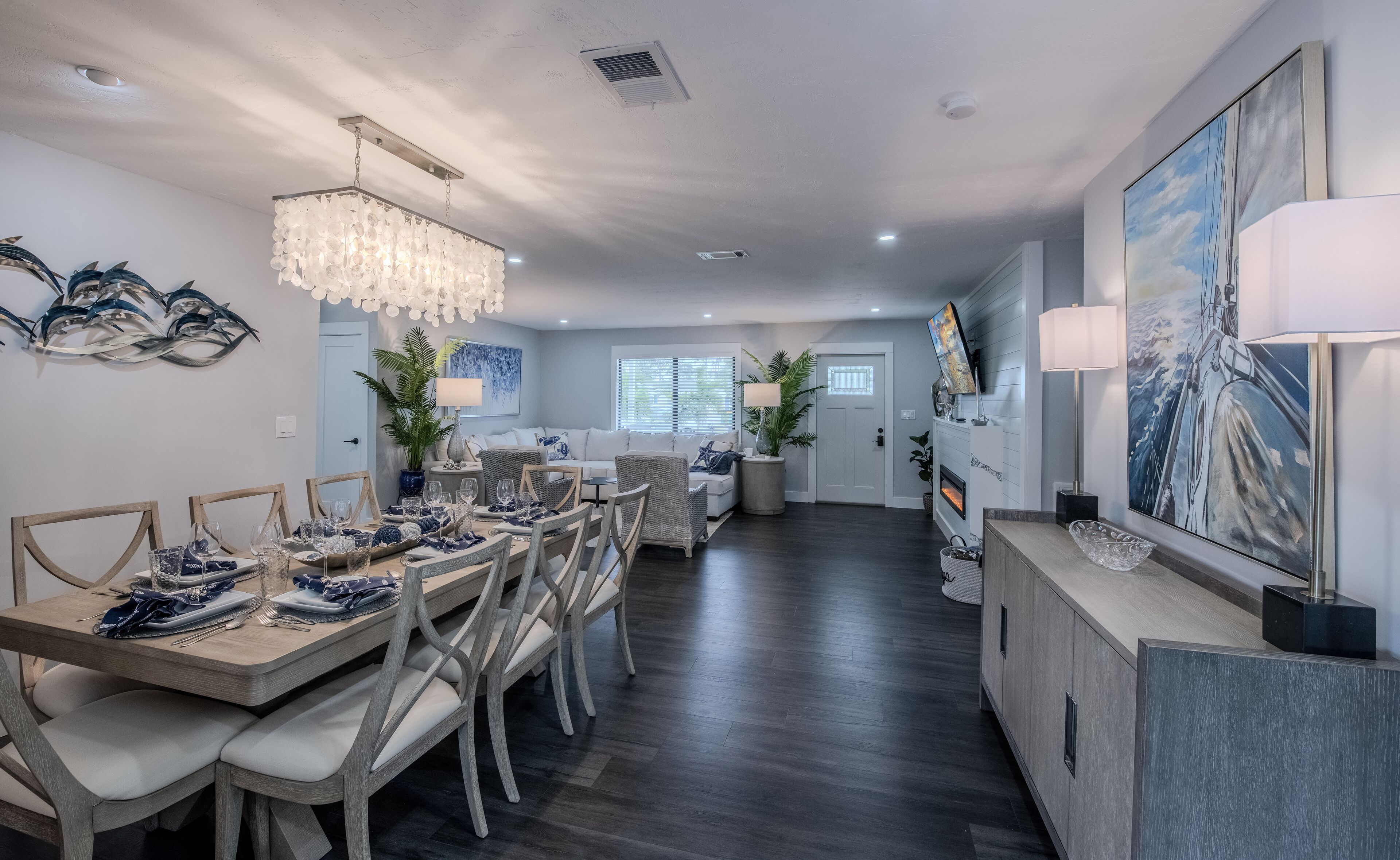 A modern dining area with a wooden table set for six, flanked by two chairs. A chandelier hangs from the ceiling, and a flat-screen TV is mounted on the wall above the fireplace.