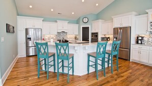A modern kitchen with white cabinets, a central island with bar stools, stainless steel appliances, and a stone backsplash. - Pa's Place, Almost Oceanfront House in Emerald Isle, NC! (Emerald Isle)