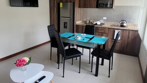A modern kitchen with dark wood cabinets, a stainless steel refrigerator, a microwave, and a granite countertop. There is a dining table with four chairs and a small white table with a vase of flowers. - Suite moderna 301. La Mejor Ubicación

 (Quito)