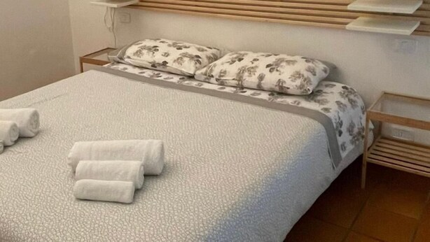 Un letto rifatto con cuscini a motivi floreali e un piumone bianco, una testiera in legno a listelli e un comodino con una lampada.