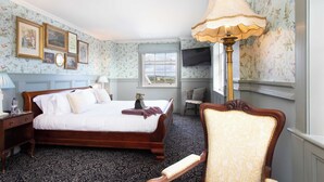 A bedroom with a large bed, a wooden nightstand, a chair, a TV, and a window with a view. - The Duchess of Cornwall Inn (Dorchester)